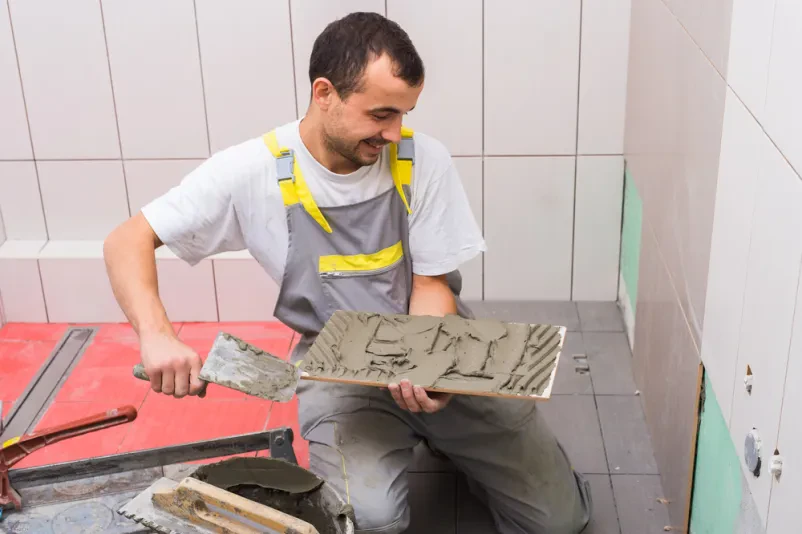 Keramičar v delovni opremi nanaša lepilno maso na ploščice in jih polaga na steno v Kočevju z okolico.