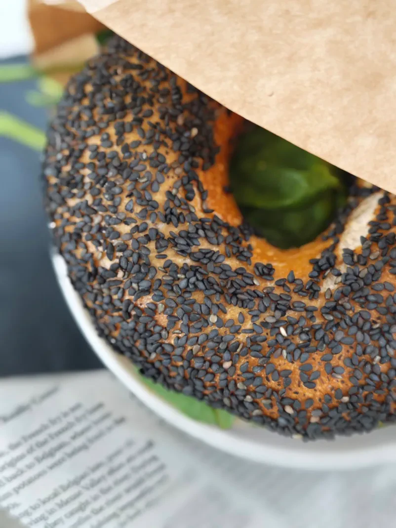 Bagel s semeni na krožniki in v papirnati vrečki iz pekarne v Ljubljani.
