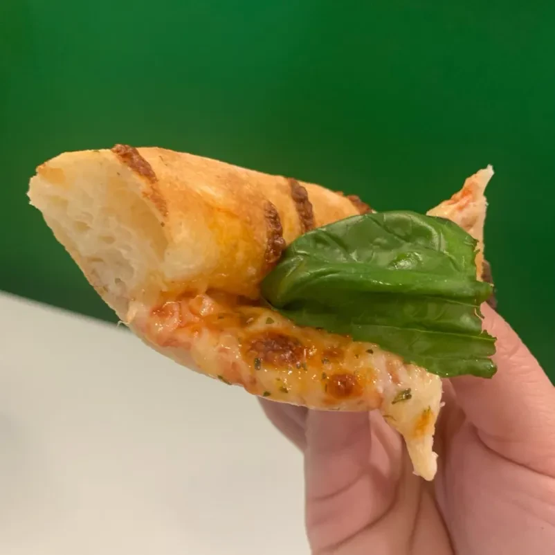Roka drži kos pizze margherite na katerem je velik list bazilike.