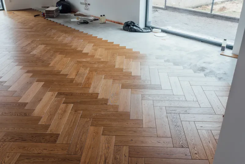 Delavec na tla stanovanjskega objekta polaga nov parket. Ob robu prostora je zložena oprema za delo in material za parketarska dela.