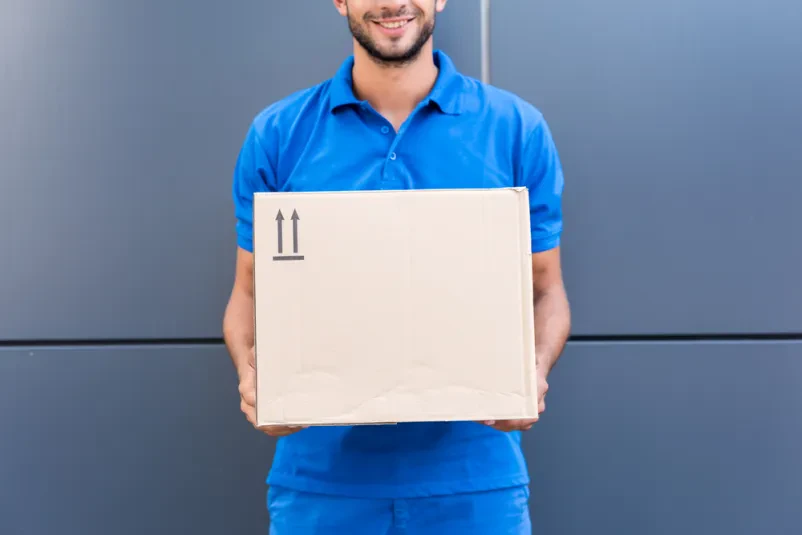 Kurir v modrih delovnih oblačilih v roki drži paket v škatli.