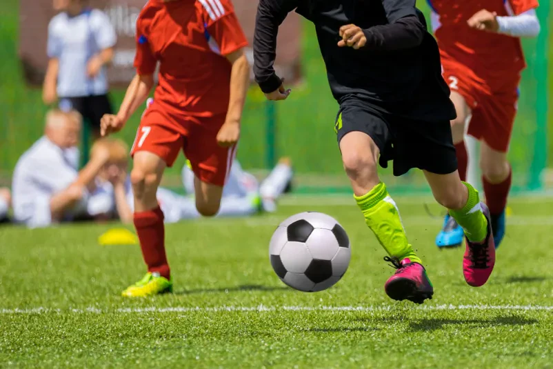 Mladinski nogometni trening na igrišču Skupina mladih nogometašev teče z žogo po sončnem zelenem nogometnem igrišču med intenzivnim treningom, ki izboljšuje njihove spretnosti in timsko delo.