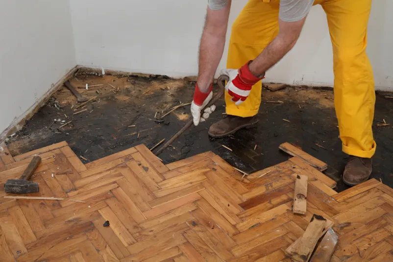 Delavec v rumenih delovnih hlačah obnavlja parket, saj odstranjuje star in obrabljen parket v Postojni.