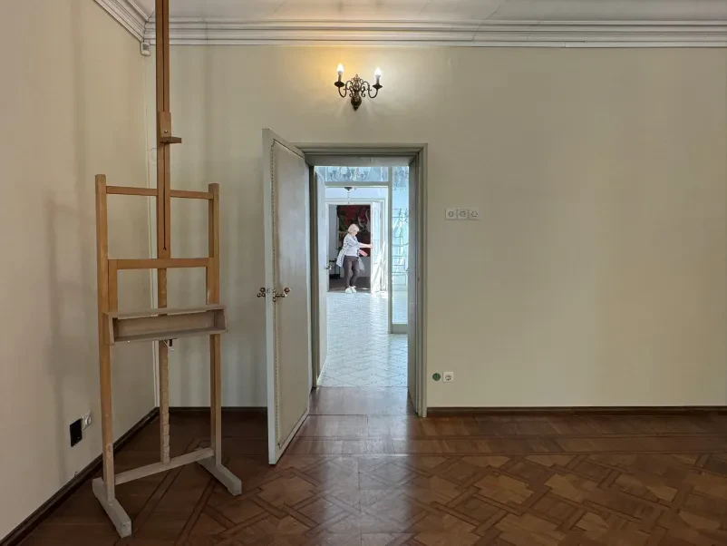 Prazno stanovanje s slikarskim platnom, v ozadju nepremičninska posrednica v sosednjem prostoru