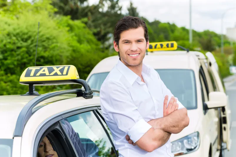 Moški voznik taksija stoji poleg vozila, ki je označen z rumeno tablo, na kateri piše Taxi.