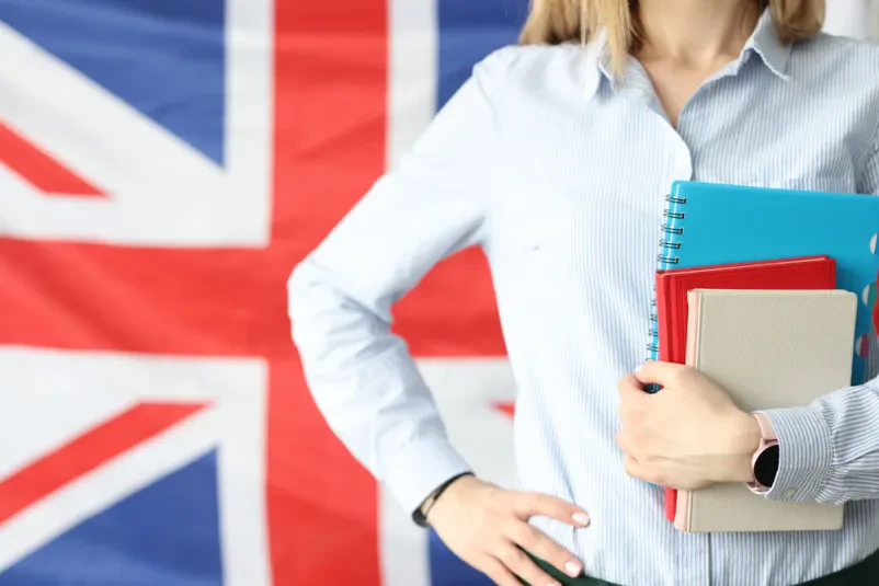 Učiteljica angleškega jezikovnega tečaja pred britansko zastavo s knjigami v rokah.