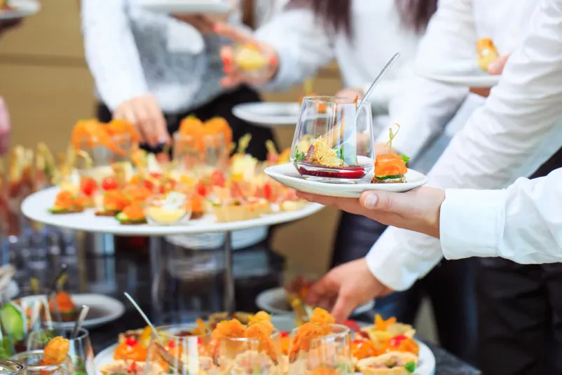 Skupina ljudi se druži in streže ob bogato obloženi mizi s catering hrano, ki je primerna za večje dogodke v Sloveniji.