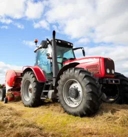 Traktor z balirko spretno zbira posušeno seno na širokem zelenem polju pod modrim nebom. To kmetijsko delo je ključno za pripravo krme.
