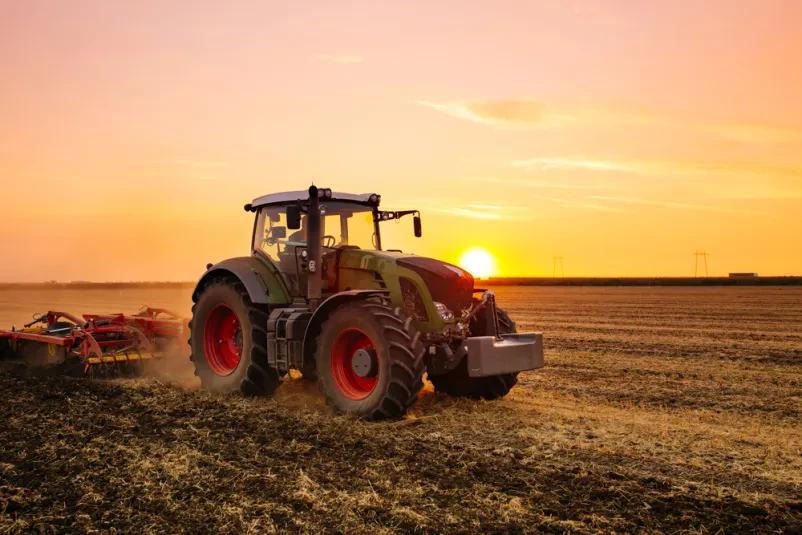 Oranje polja s traktorjem ob sončnem zahodu Rdeč traktor v večerni svetlobi orje njivo, dviguje prst za setev. To so tipična dela s traktorjem v Mavčičah, Kranju in okolici Gorenjske.