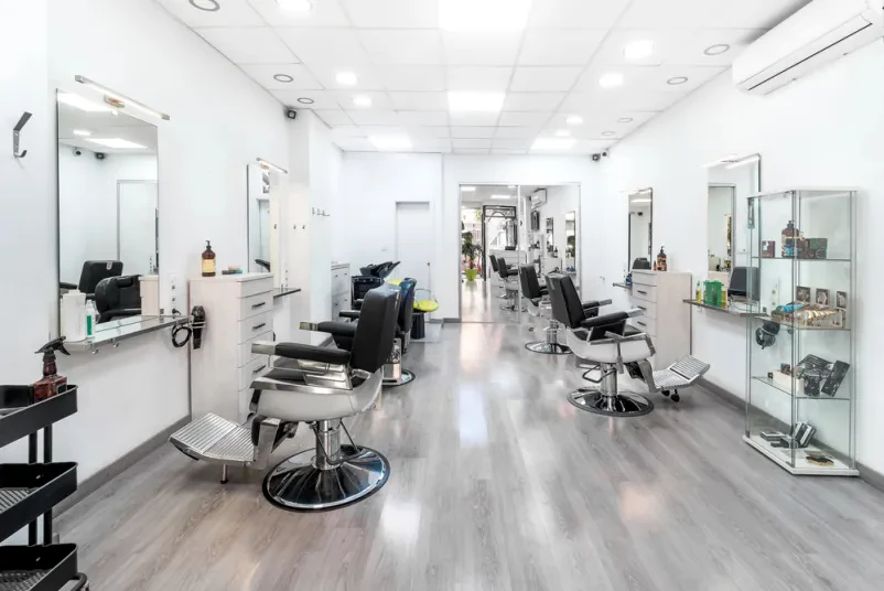 Svetel frizerski salon z modernim črno-belim dizajnom. Vidni so ogledala in stoli za stranke, ki ustvarjajo urejeno in vabljivo okolje.