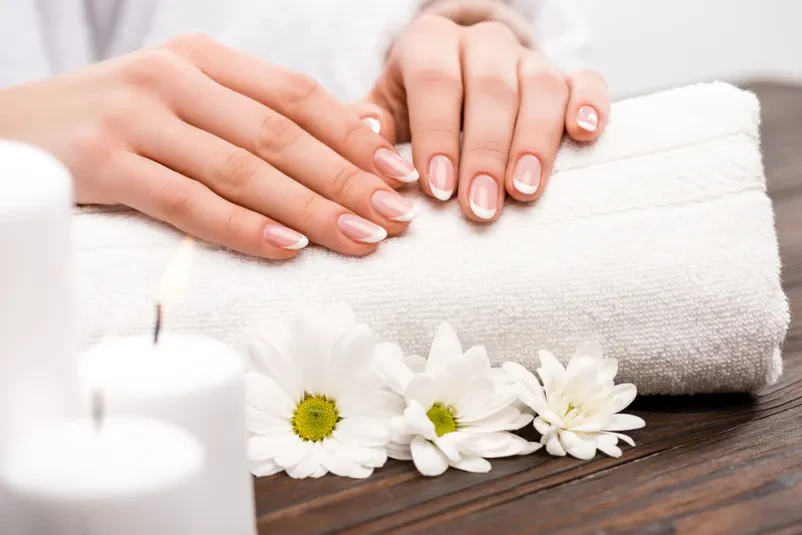 Bližnji posnetek ženske roke med sproščujočo spa nego, obkrožene z nežnimi rožami in dišečimi svečami za popolno manikuro in dobro počutje.