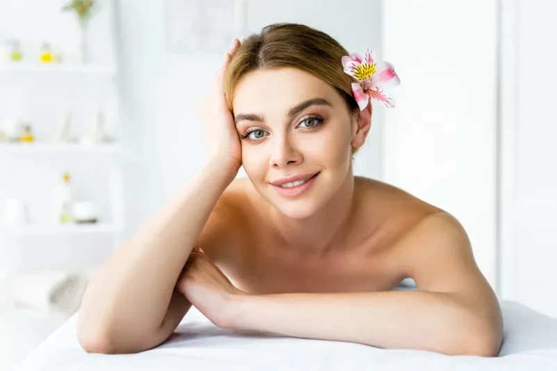 Nasmejana ženska sproščeno leži na masažni mizi, z belim cvetom na hrbtu. Prizor nakazuje globoko sprostitev in dobro počutje med wellness tretmajem.