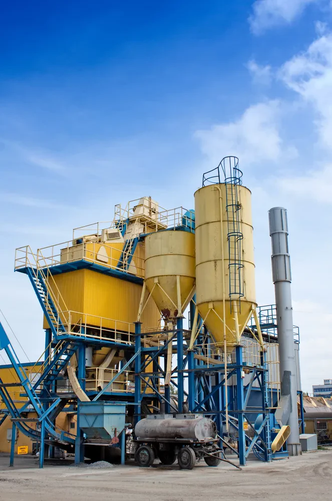 Tovarna cementa z velikimi rumenimi silosi Industrijski kompleks tovarne cementa s poudarjenimi rumenimi silosi, ki se dvigajo proti nebu. Objekt je opremljen za pripravo različnih betonskih mešanic in izdelavo betona.
