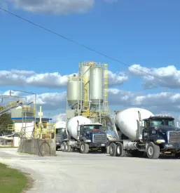 Panoramski posnetek obrata za proizvodnjo betona z velikimi rezervoarji in skladiščnimi prostori. Industrijski kompleks je postavljen pod jasnim modrim nebom.