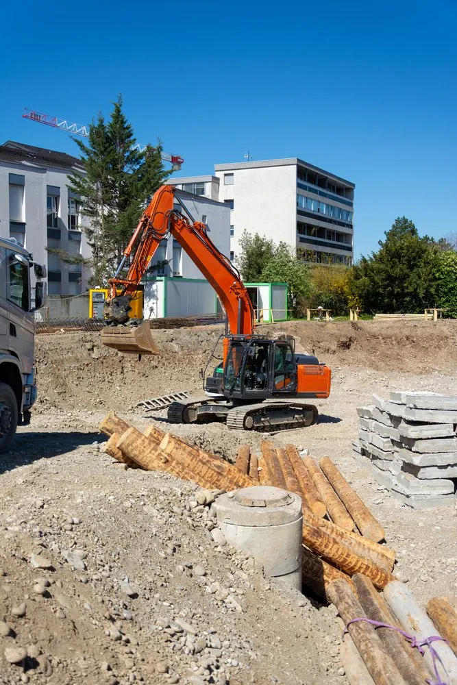 Gosenični bager v globoki gradbeni jami, obdan z lesenimi piloti in betonskimi ploščami, izvaja izkope z bagrom Celje.