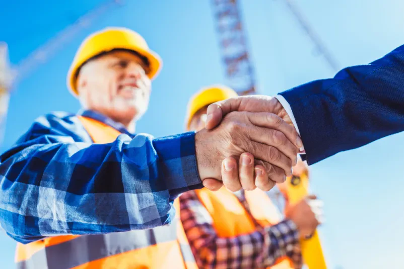 Gradbenik in poslovnež si segata v roke Gradbeni delavec v zaščitni opremi in poslovnež v elegantni obleki si segata v roke na gradbišču, kar simbolizira uspešno predajo projekta ali sklenitev pogodbe za gradnjo objektov na ključ.