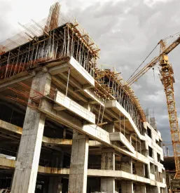 Visok rdeč žerjav stoji poleg nedokončane večnadstropne stavbe, ki se dviga proti oblačnemu nebu, kar kaže na intenzivno fazo gradnje novih objektov v urbanem okolju.