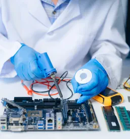 Elektronik se osredotoča na delo s komponentami, uporablja voltmeter za testiranje in preverjanje vezij v specializirani delavnici. Miza je polna orodja in opreme.