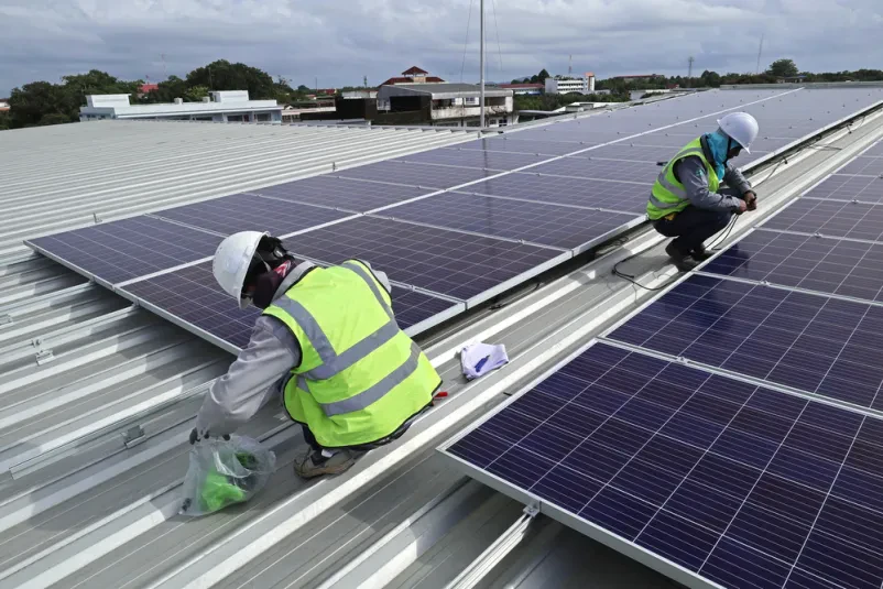 Tehnik povezuje kable solarnega sistema Tehnik v zaščitni čeladi skrbno povezuje kable na strehi industrijske stavbe. To zagotavlja optimalno in varno delovanje nameščenih solarnih modulov.