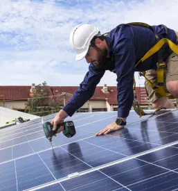 Tehnik v delovni uniformi z električnim vrtalnikom pritrjuje sončne kolektorje na streho. S tem zagotavlja stabilnost in dolgotrajnost solarne instalacije.