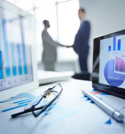 Očala, pisalo, dve tablici in finančni dokumenti na delovni mizi, v ozadju pa se poslovneži dogovarjajo. Nudimo pomoč pri poslovanju in optimizacijo poslovanja.