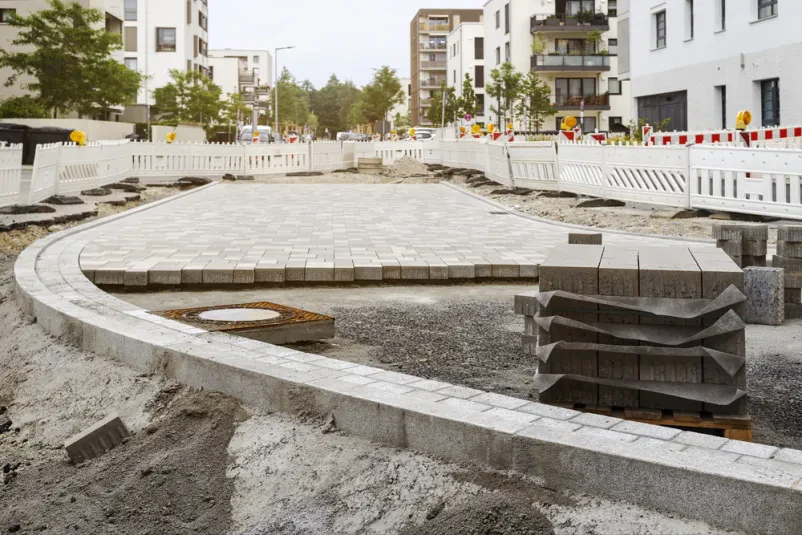 Polaganje tlakovcev pri cestnih delih Prikaz del na gradbišču, kjer se polagajo tlakovci za prenovo cestne površine. Delo poteka ob zaobljenem modernem odseku ceste, kar kaže na strokovno tlakovanje poti.