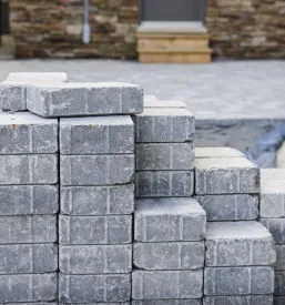 Veliki kupi sivih prepletenih tlakovcev, pripravljenih za uporabo pri urejanju okolice. Ti bloki so idealni za vzdržljivo tlakovanje dovoza in drugih zunanjih površin.