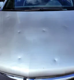 Bližnji posnetek pokrova motorja osebnega vozila, ki kaže jasno vidne udrtine, povzročene s točo. Poškodbe so razpršene po celotni površini haube in zahtevajo kleparsko popravilo.