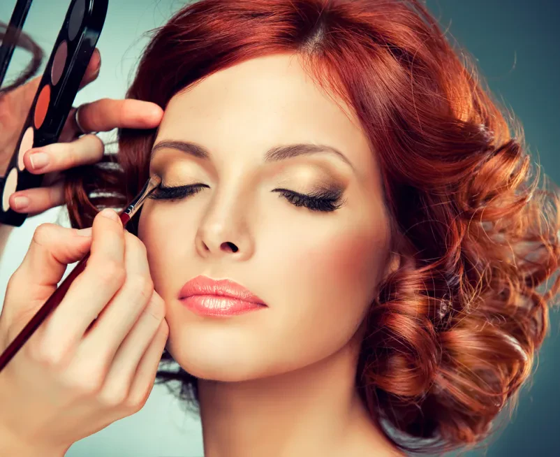 Vizažistka skrbno liči manekenko za glamurozen videz Umetnica ličenja, osredotočena na delo, nanaša glamurozen make up rdečelasi manekenki za modno snemanje.