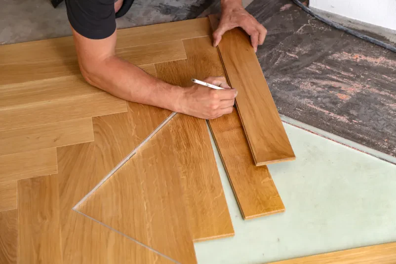 Delavec namešča lesene talne obloge Delavec previdno namešča leseno laminatno ploščo na tla, s poudarkom na natančnosti in pravilni postavitvi.