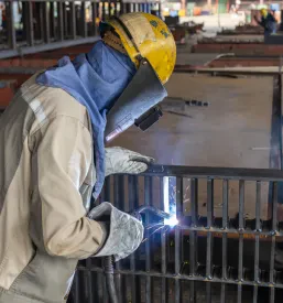 Varilec v celotni zaščitni opremi skrbno vari kompleksno jekleno konstrukcijo v industrijskem okolju, zagotavljajoč varnost in natančnost.