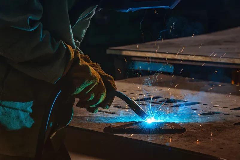 Varilec kovin med delom Delavec, opremljen z zaščitno masko, zvarja kovino v industrijski hali, pri čemer ustvarja svetle iskre. To je klasičen primer varilstva.