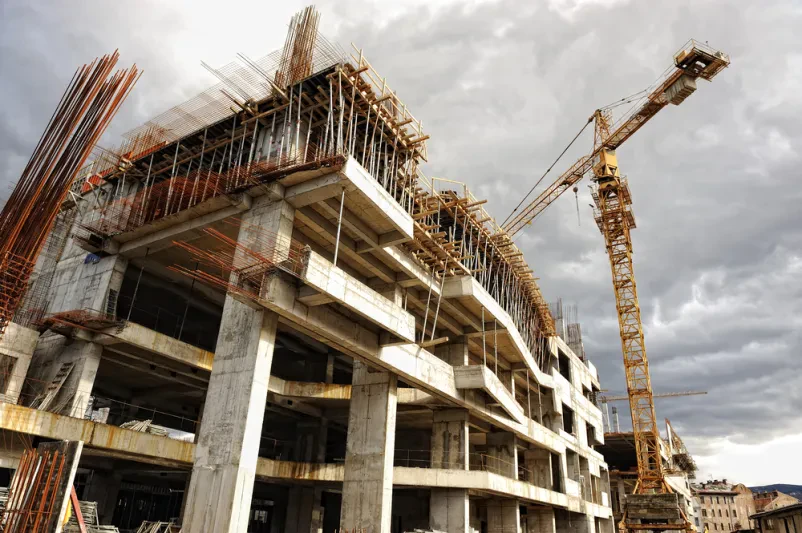 Visok rdeč gradbeni žerjav se dviga nad nedokončano konstrukcijo moderne stavbe, poudarjajoč razvoj in napredek v gradbeništvu.