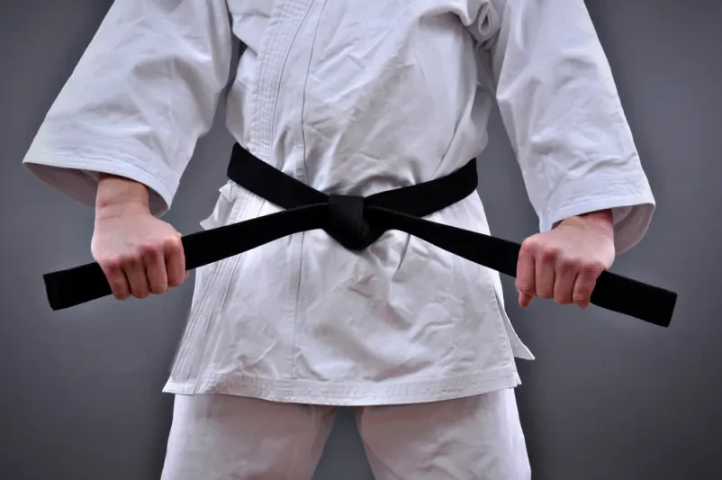 Moški v uniformi borilnih veščin ponosno drži svoj črni pas z obema rokama, simbol discipline in dolgoletnih dosežkov.