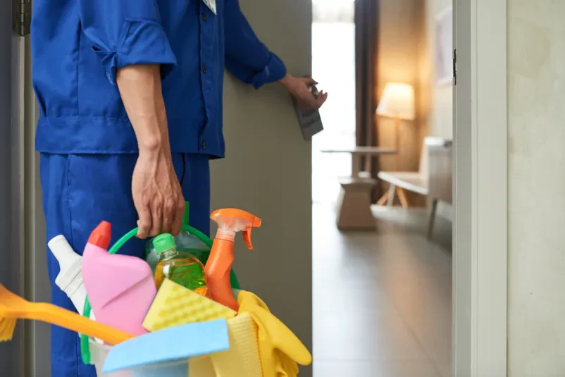 Profesionalni čistilec vstopa v sobo s čistilnimi sredstvi Profesionalni čistilec z vedrom čistilnih sredstev vstopa v svetlo sobo. Pripravljen je za temeljito čiščenje nastanitev.