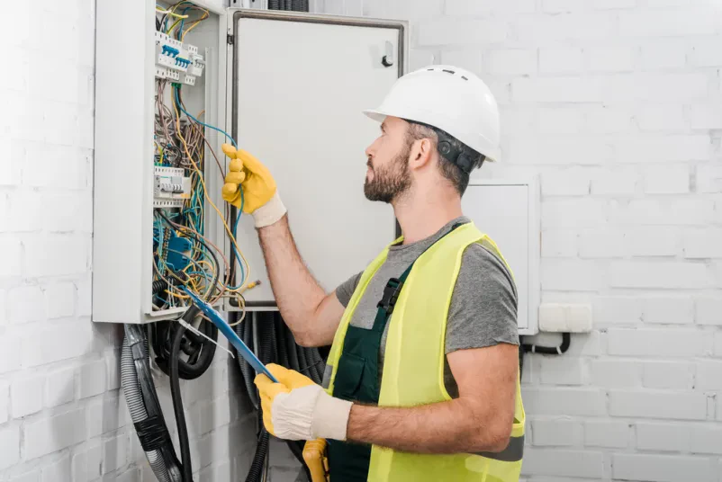 Električar preverja žice v omarici Stranski pogled na električarja v rumenem jopiču, ki s pomočjo sponke in beležke skrbno pregleduje in preverja žice v električni omarici na hodniku.