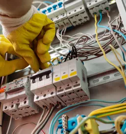 Električar v zaščitnih rokavicah, posvečen svojemu delu, skrbno popravlja in namešča komponente v električni razdelilni omarici. Poudarek na varnosti.