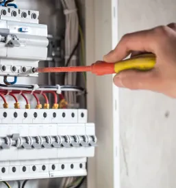 Elektro tehnik z orodjem v rokah pozorno dela v stikalni plošči, kjer pregleduje in povezuje električne varovalke za pravilno delovanje sistema.