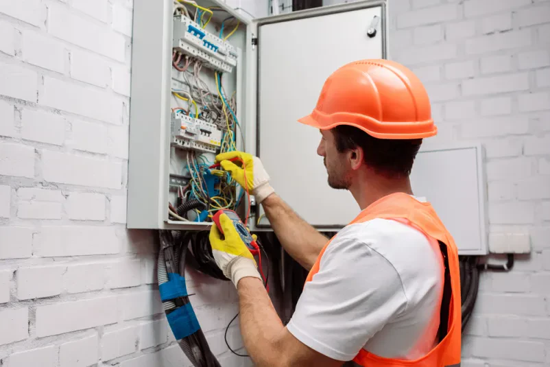 Električar z multimetrom ob razdelilni omarici Usposobljen električar v zaščitni opremi pregleduje električno razdelilno omarico z multimetrom. Poiščite dober električar Zasavje za varno in zanesljivo storitev.