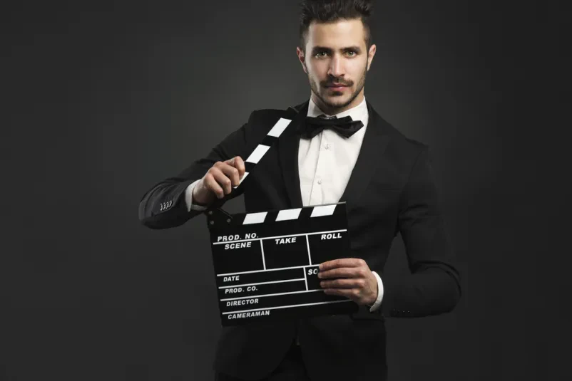 Moški drži filmsko klapo Portret elegantnega moškega v smokingu, ki drži filmsko klapo. Slika poudarja profesionalnost v filmski industriji.
