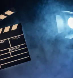 Roka operaterja drži filmsko klapo, v ozadju pa je zamegljena studijska luč. Prizor nakazuje potek filmske produkcije.