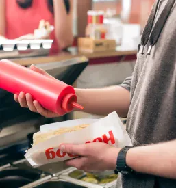 Prijazen prodajalec s svežimi sestavinami skrbno pripravlja hotdog v okrepčevalnici. To kakovostno in hitro storitev nudi dobra okrepčevalnica Murska Sobota.