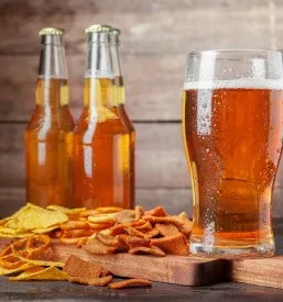 Kozarec svetlega piva lager in različni slani prigrizki, vključno z oreščki, čipsom in prestami, privlačno razporejeni na leseni mizi.