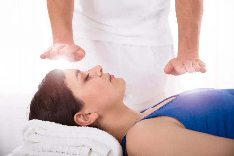 Bližnji posnetek sproščene mlade ženske med reiki zdravljenjem, kjer roke terapevta nežno delujejo na območju obraza.