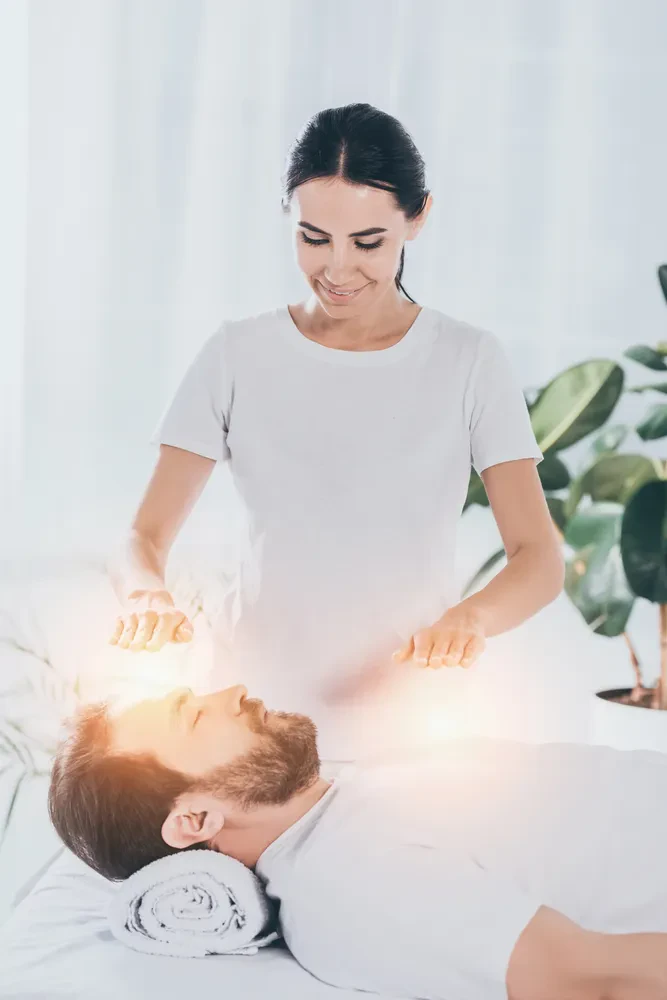 Nasmejana mlada terapevtka izvaja reiki seanso za bradatega moškega z zaprtimi očmi, ki leži sproščen na terapiji.
