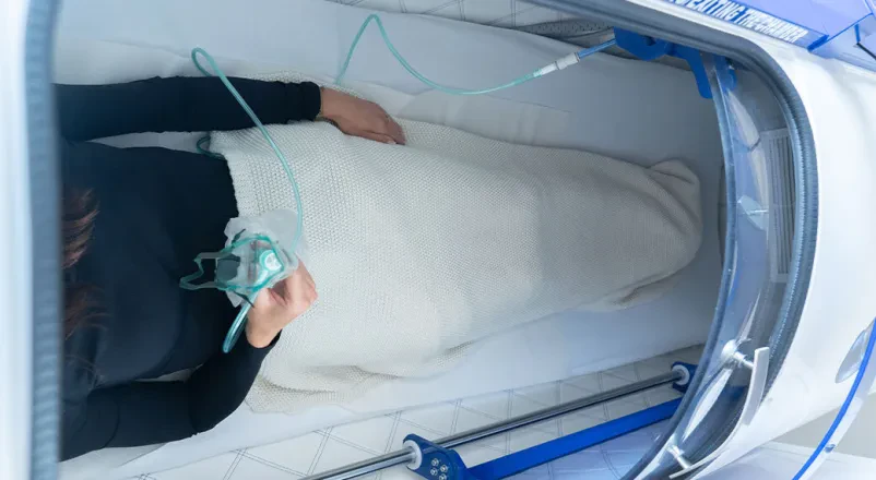 Uporaba kisikove komore Posameznik leži v kisikovi komori, opremljen z masko za vdihovanje čistega kisika, za podporo naravnim procesom celjenja.