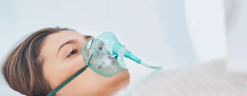 Hiperbarična terapija z masko za kisik Oseba sproščeno leži v hiperbarični komori in diha kisik skozi masko, kar pomaga pri Hiperbarična kisikova terapija Kamnik, okolica.