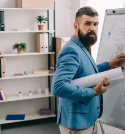 Zrelejši moški arhitekt v elegantni modri obleki drži načrt in razlaga pred flip chart tablo v pisarni. Za inženiring in tehnično svetovanje Krško, Posavje smo vam na voljo.