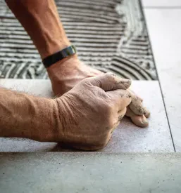 Roke strokovnjaka polagajo velike keramične ploščice na betonska tla. Uporabljeno je kakovostno lepilo za optimalen in dolgotrajen oprijem.