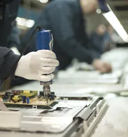 Delavci natančno sestavljajo elektronske komponente na proizvodni liniji v zaprtem prostoru, kar ponazarja industrijsko elektromontažo in delo s šibkim tokom.